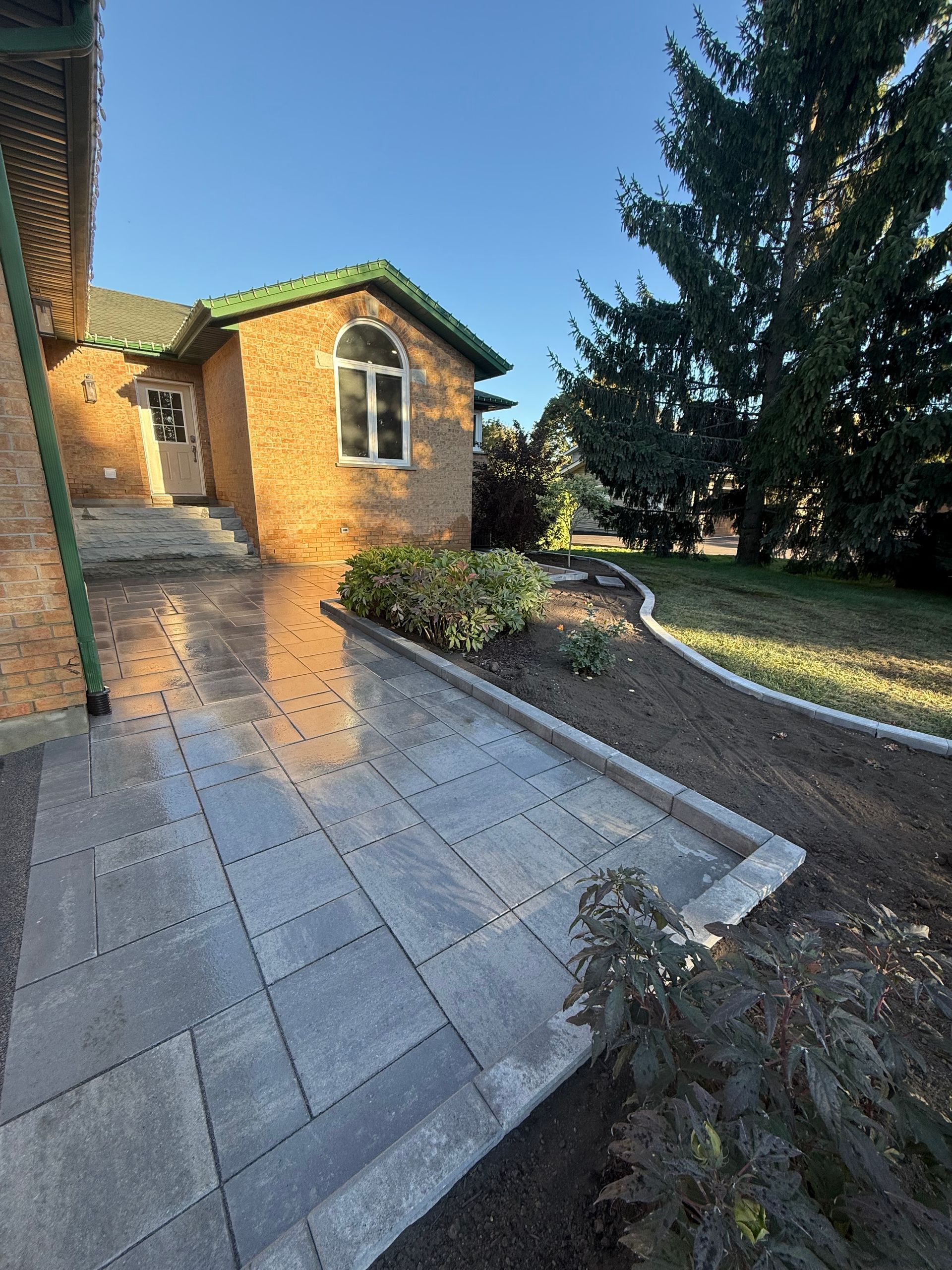 Gray stone walkway leading to a tan house with green roof, landscaped garden and tree in the background.