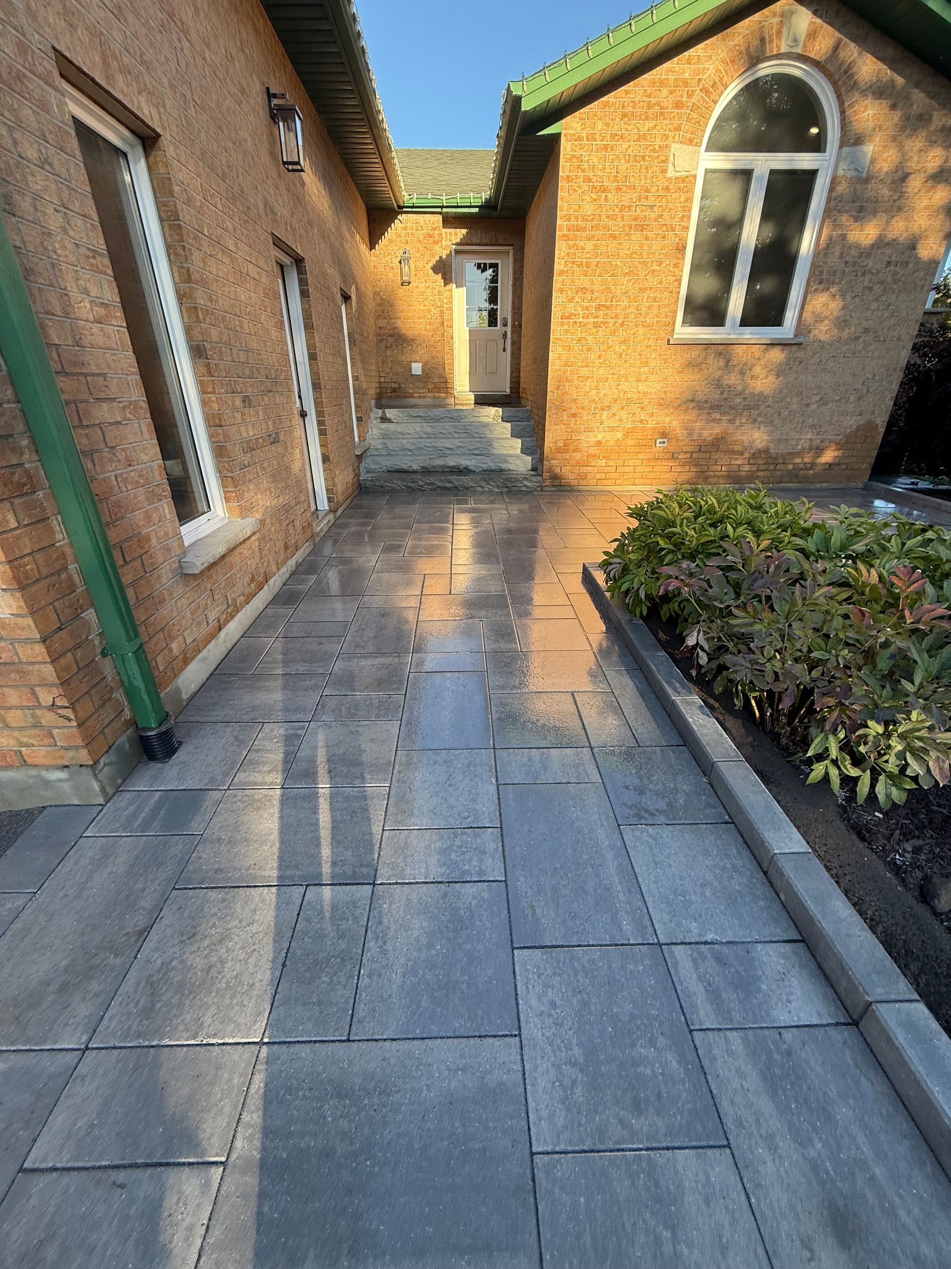 Brick buildings flank a gray paved walkway leading to a doorway. Green foliage and a gutter are visible.
