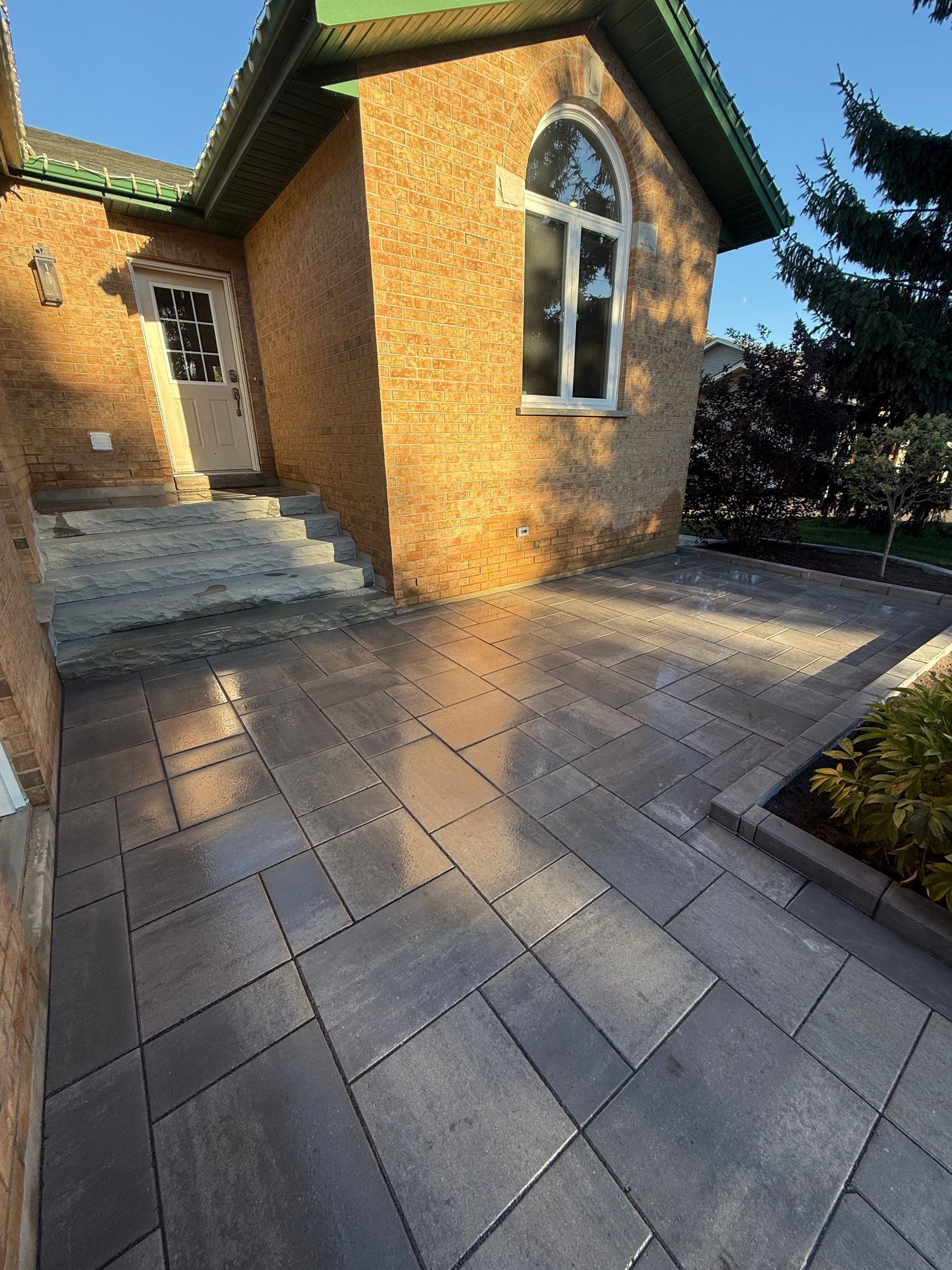Brick house entrance with gray paved walkway. Arched window, steps, and green roof.