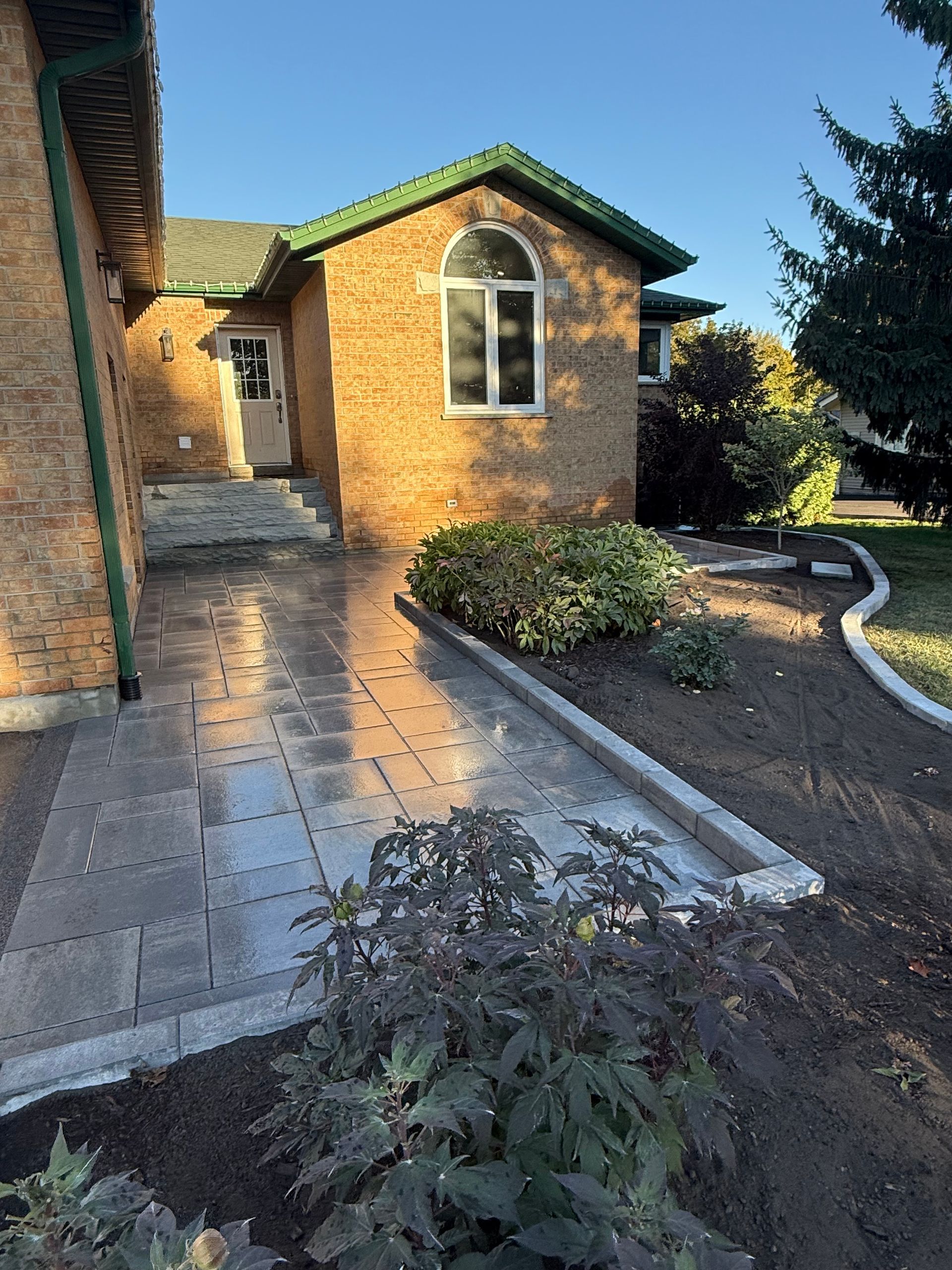 Brick house with paved walkway, flower beds, and a green roof under a blue sky.
