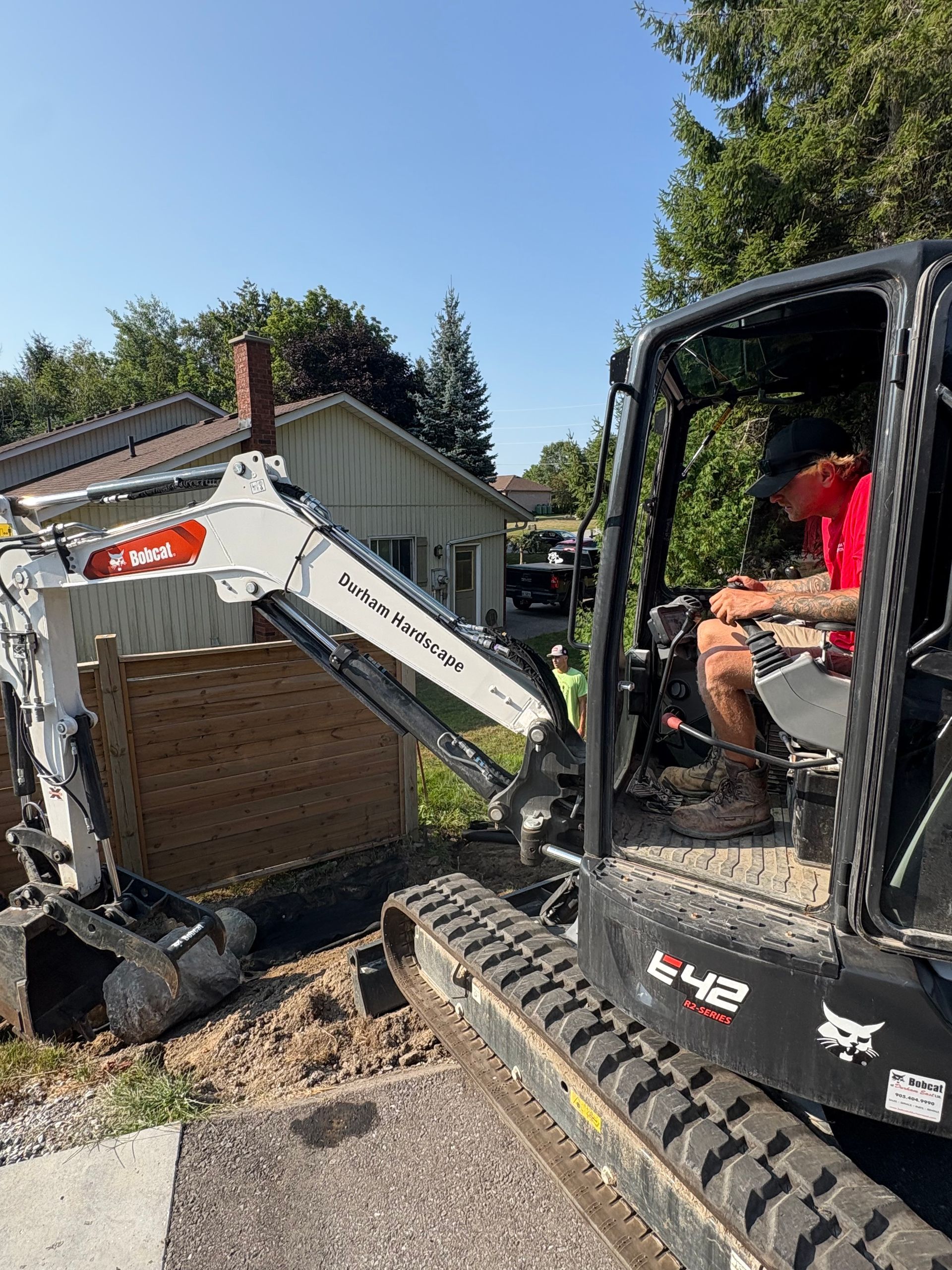 A Bobcat excavator digs near a house, operated by a person in a red shirt. Sunny day.