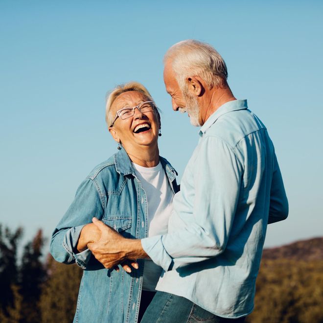 An elderly couple is laughing and holding hands in a field.