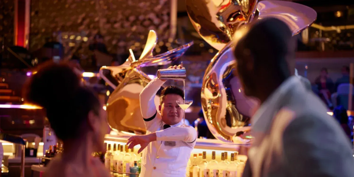 Bartender in white pours a cocktail beneath a golden dragon sculpture, with audience silhouettes in foreground