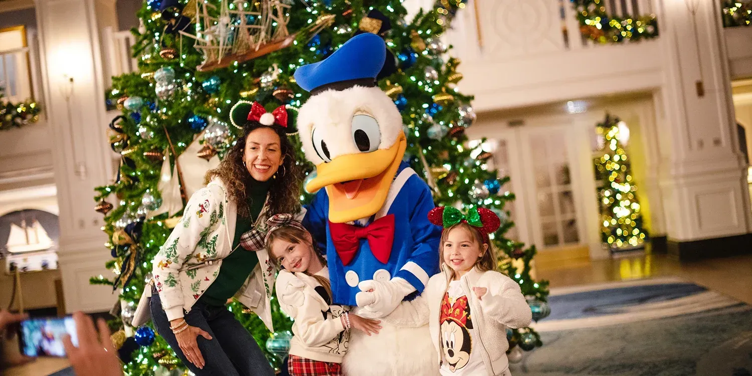 Donald Duck poses with a woman and two children by a Christmas tree in a decorated lobby.