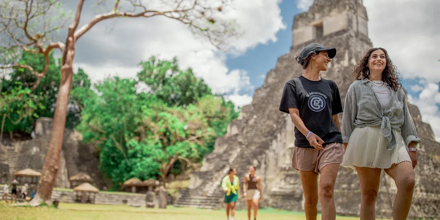 Two people walk toward camera; ancient temple in background, sunny day.