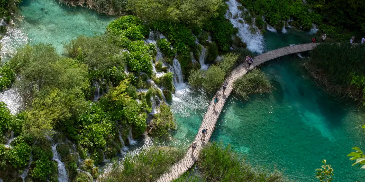 Aerial view of a wooden walkway over turquoise water beside lush green forest and a small waterfall