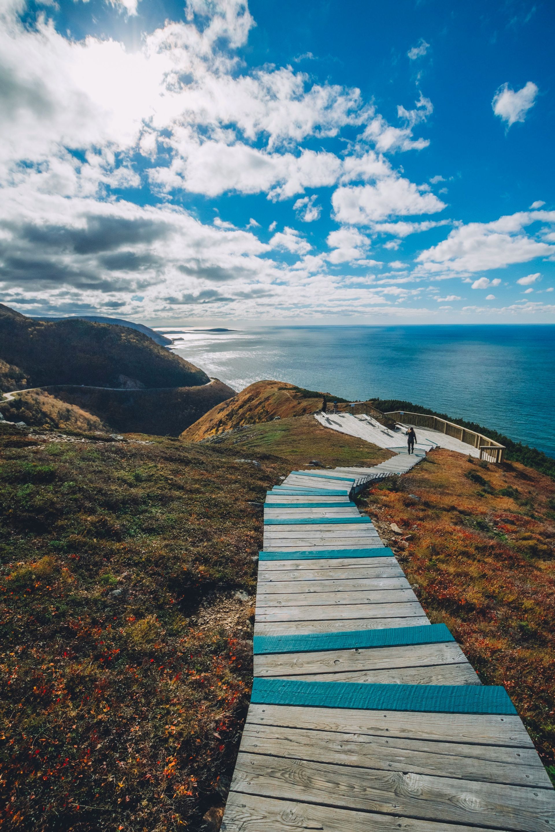 A downward-sloping wooden walkway with teal accents leads toward a sunny coastal cliff overlooking a vast blue ocean.