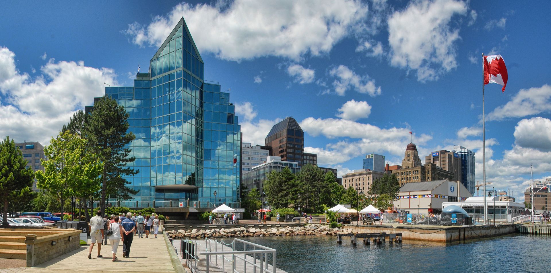 A bright city waterfront featuring a modern blue glass skyscraper, a Canadian flag, and pedestrians on a harbor walkway.