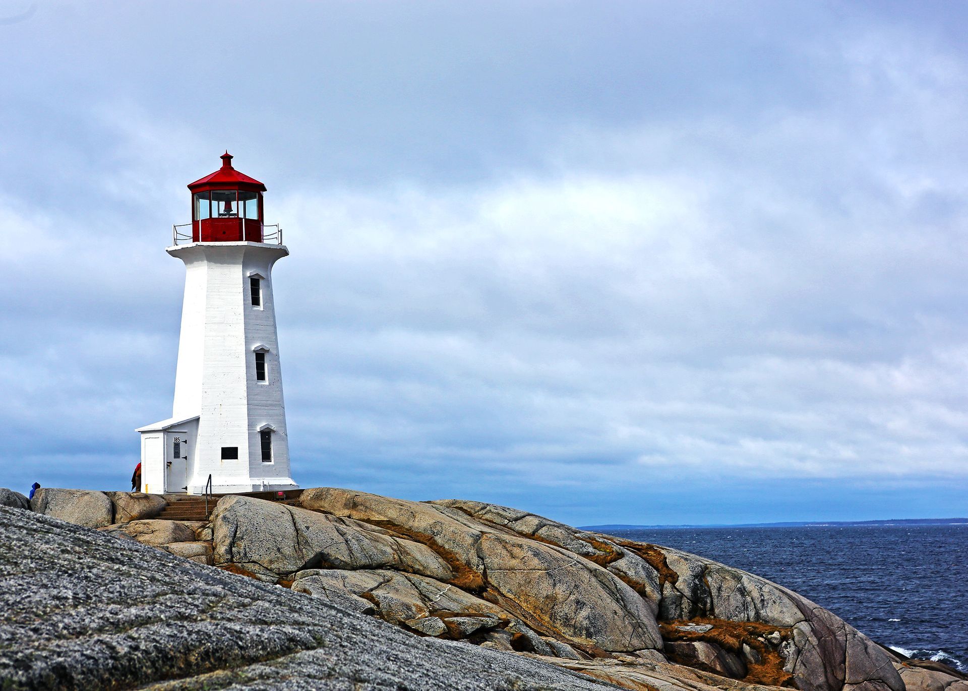 A white lighthouse with a red lantern room, perched on rocky cliffs overlooking the ocean under a cloudy sky.
