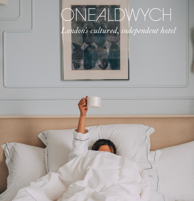 Person in bed raising a mug, light-filled hotel room. Soft linens, framed art, and a light-blue wall.