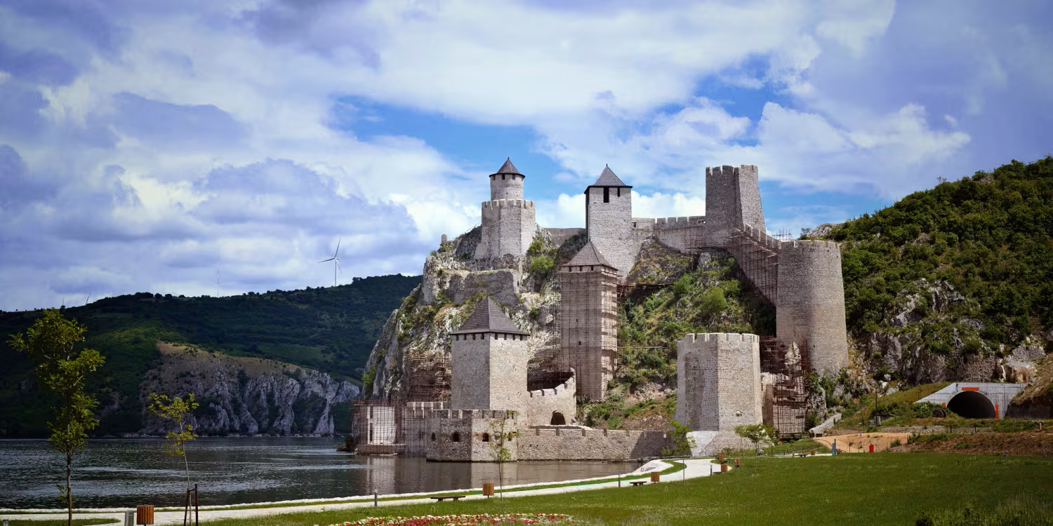 Stone castle ruin by a calm river, framed by green hills and a cloudy blue sky