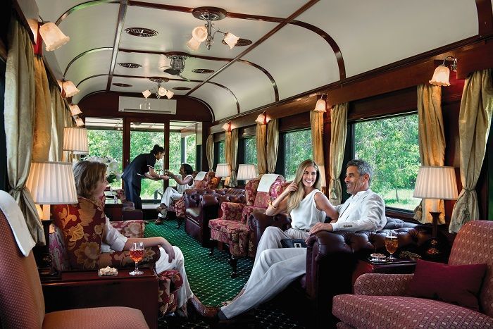 Interior of a luxury train car with people seated, a waiter, and ornate decor.