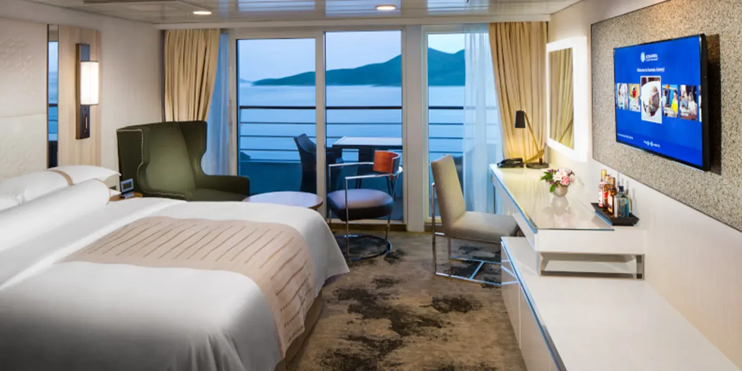 A cruise ship cabin with a bed, seating area, balcony, and a view of the water and shore.