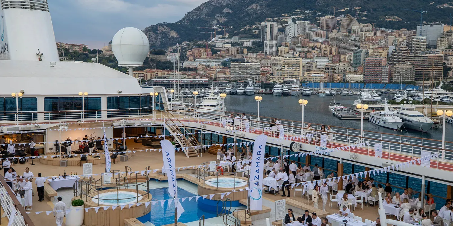 Cruise ship deck with outdoor dining, Monaco harbor in background. People at tables, yachts docked, mountains.