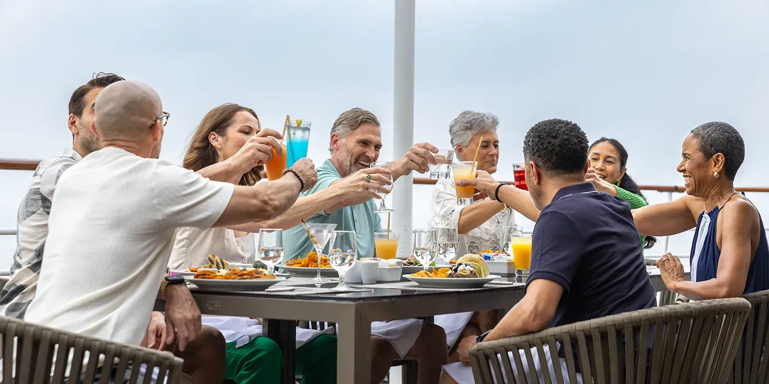 People clinking glasses at outdoor table, drinks and food present, against cloudy sky background.