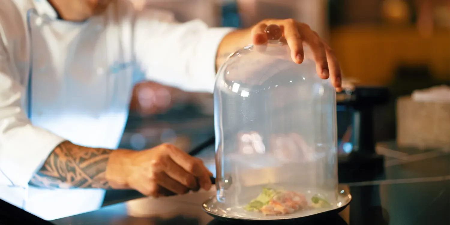 Chef placing a glass dome over a plate with food, filling the dome with smoke in a kitchen.