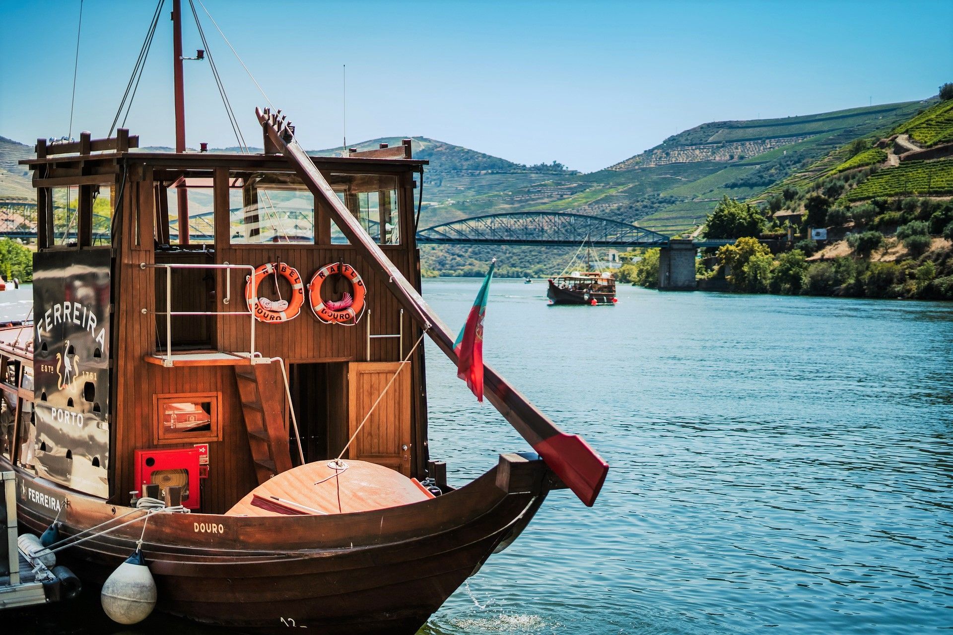 A wooden Rabelo boat with a Portuguese flag docked on the Douro River, with vineyard-covered hills in the background.
