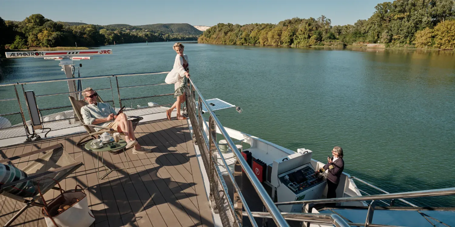 People relaxing on a boat deck, navigating a river surrounded by lush trees.