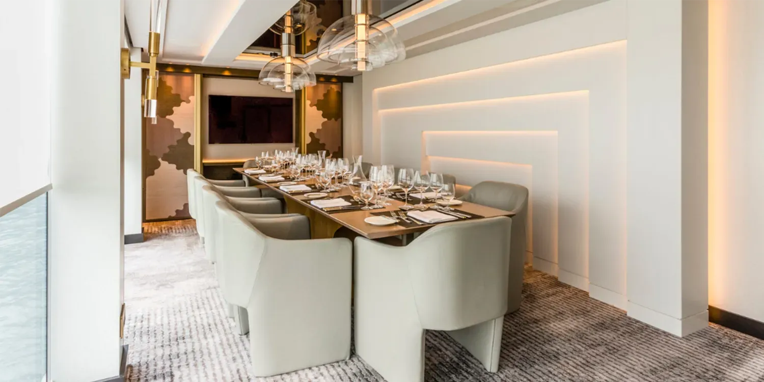 Elegant dining room with a long table set for a meal.  White walls and chairs, patterned carpet, and decorative lighting.