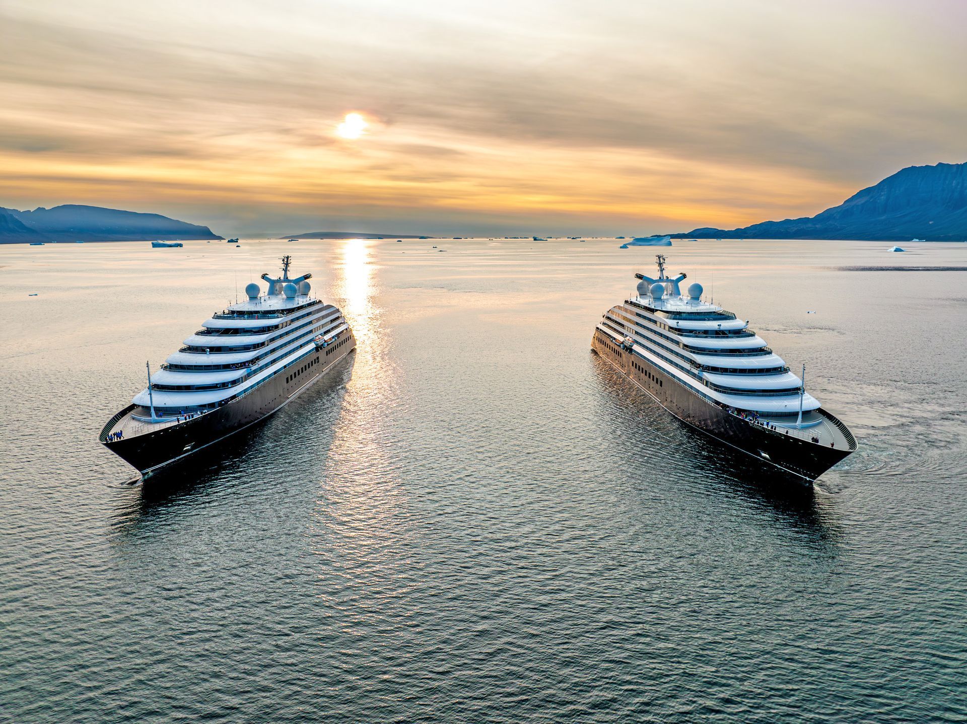 Two matching, modern cruise ships anchored side-by-side in calm, sunlit waters at dusk.