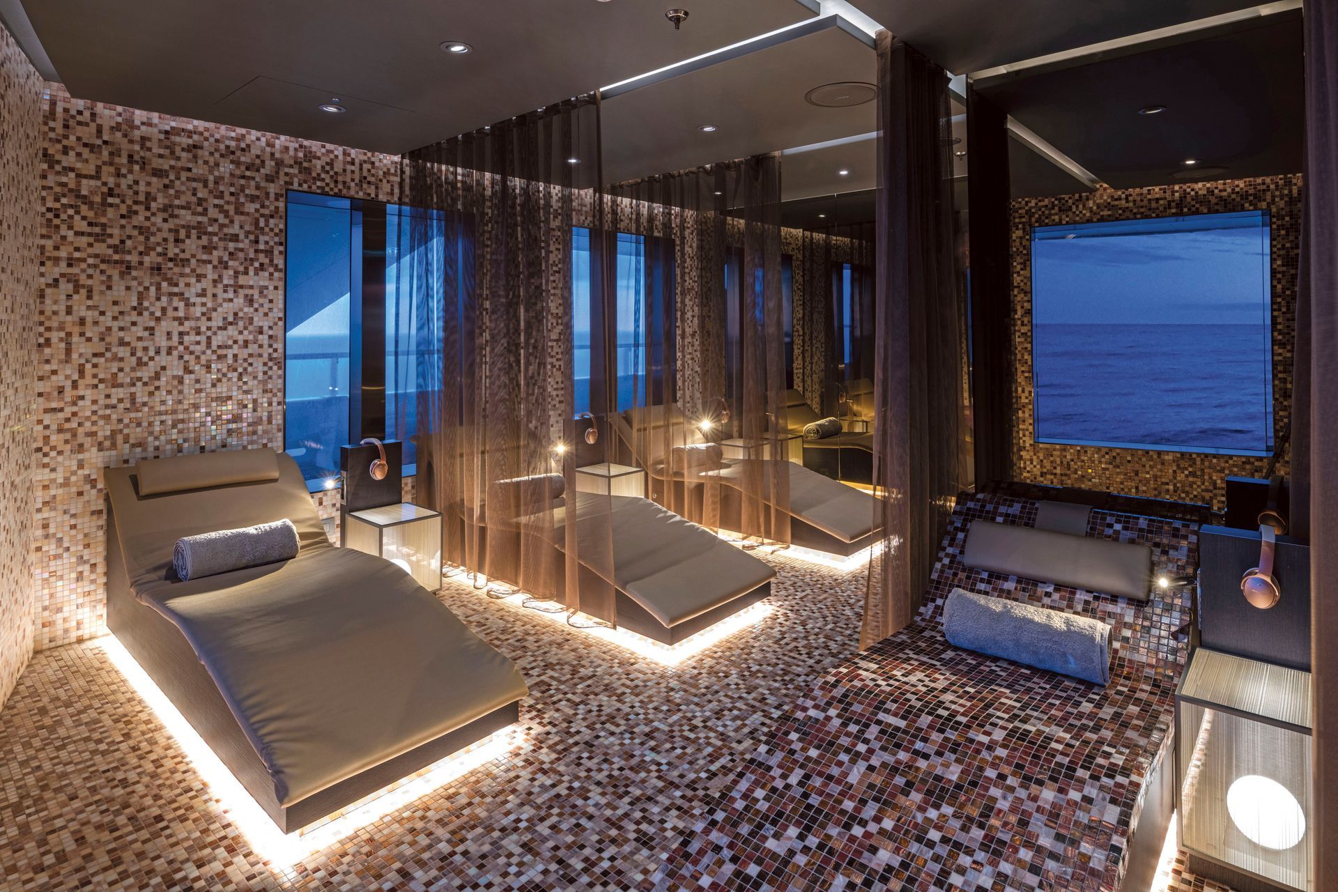 A luxury spa relaxation room with illuminated loungers, textured walls, sheer privacy curtains, and a view of the sea.