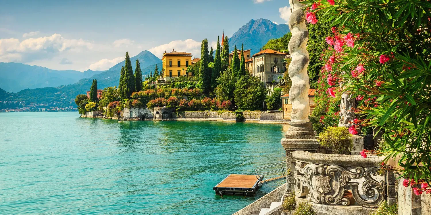 Scenic view of a lake with buildings along the shore, mountains in the background, and vibrant flowers in the foreground.