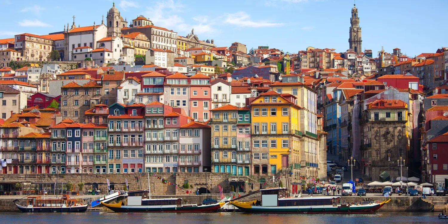 Colorful buildings line a waterfront in Porto, Portugal, with boats on the water and a tall tower in the background.