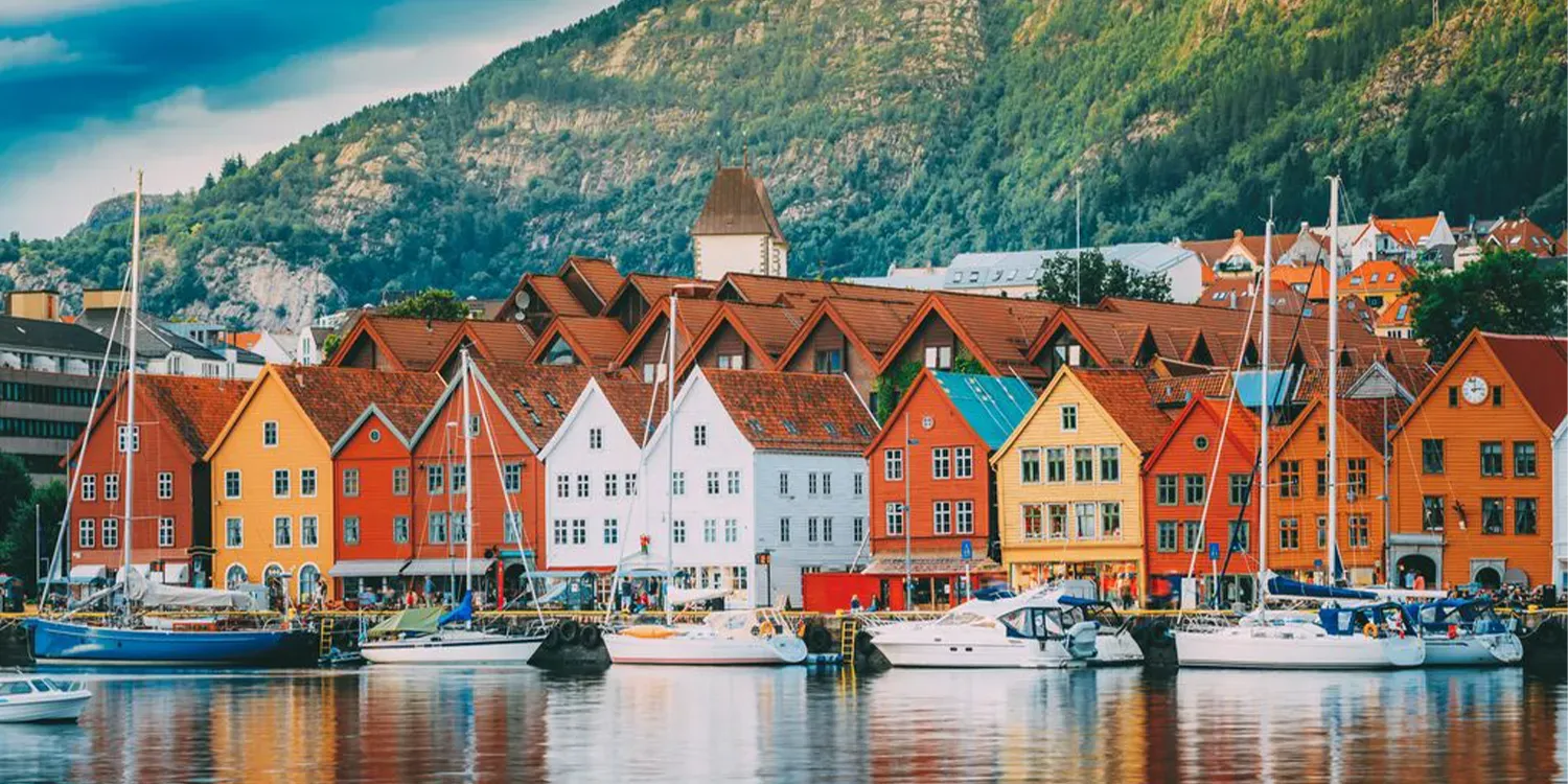 Colorful wooden buildings line a harbor with boats, mountains in background.