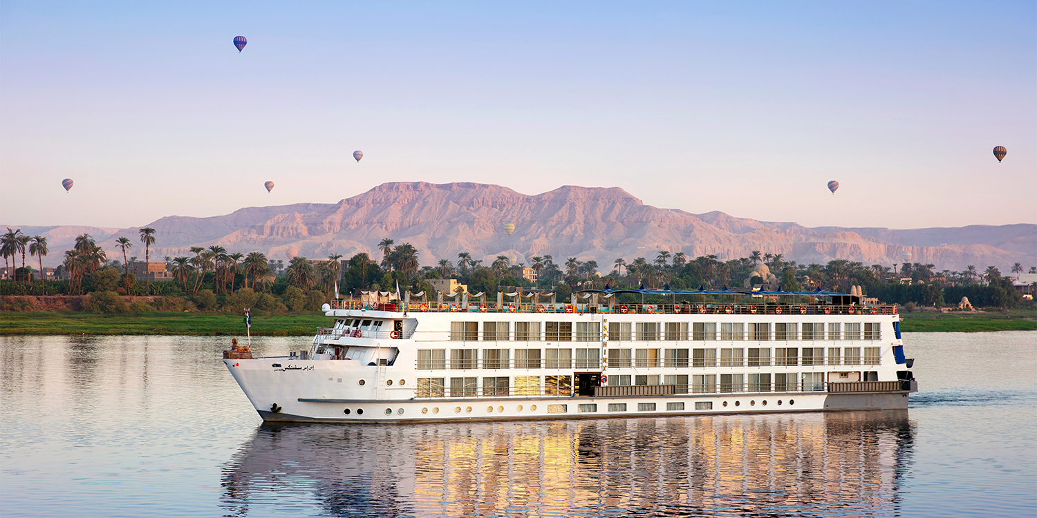 Cruise ship sailing on a river, with mountains and hot air balloons in the background.