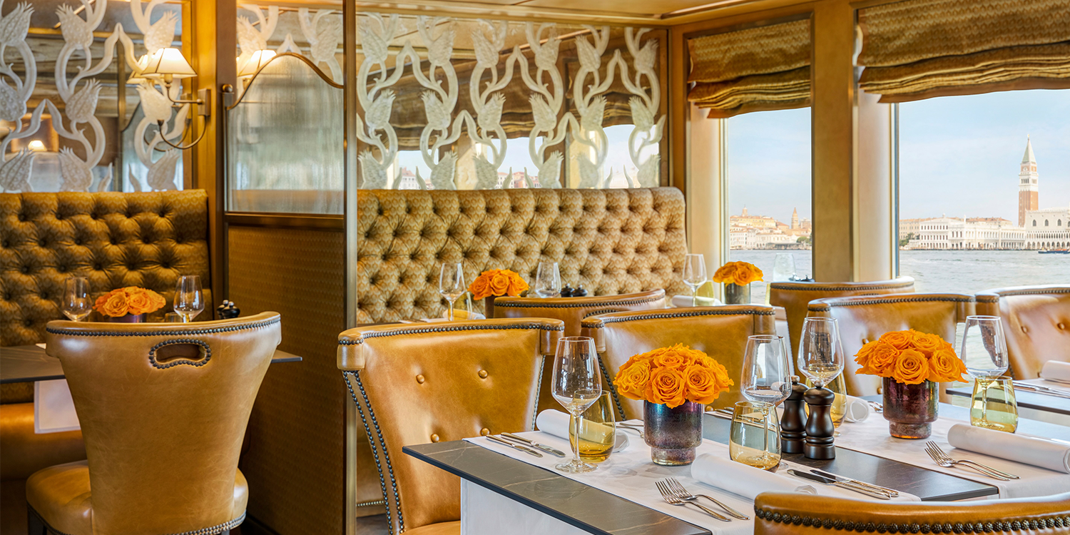 Elegant restaurant interior with tables set for dining, Venetian cityscape visible through a window.