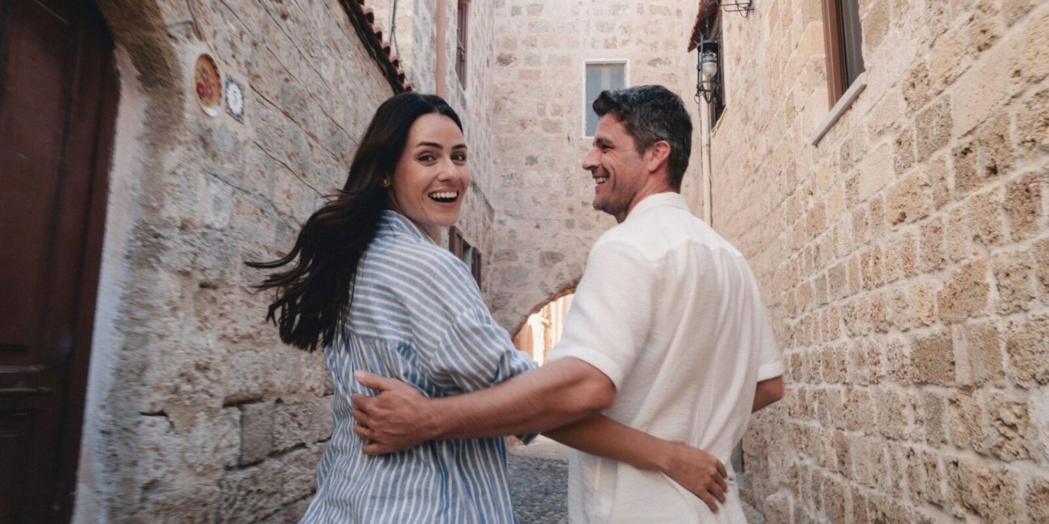 A couple smiles while walking in a stone-walled alley. The woman looks back, while the man keeps forward.