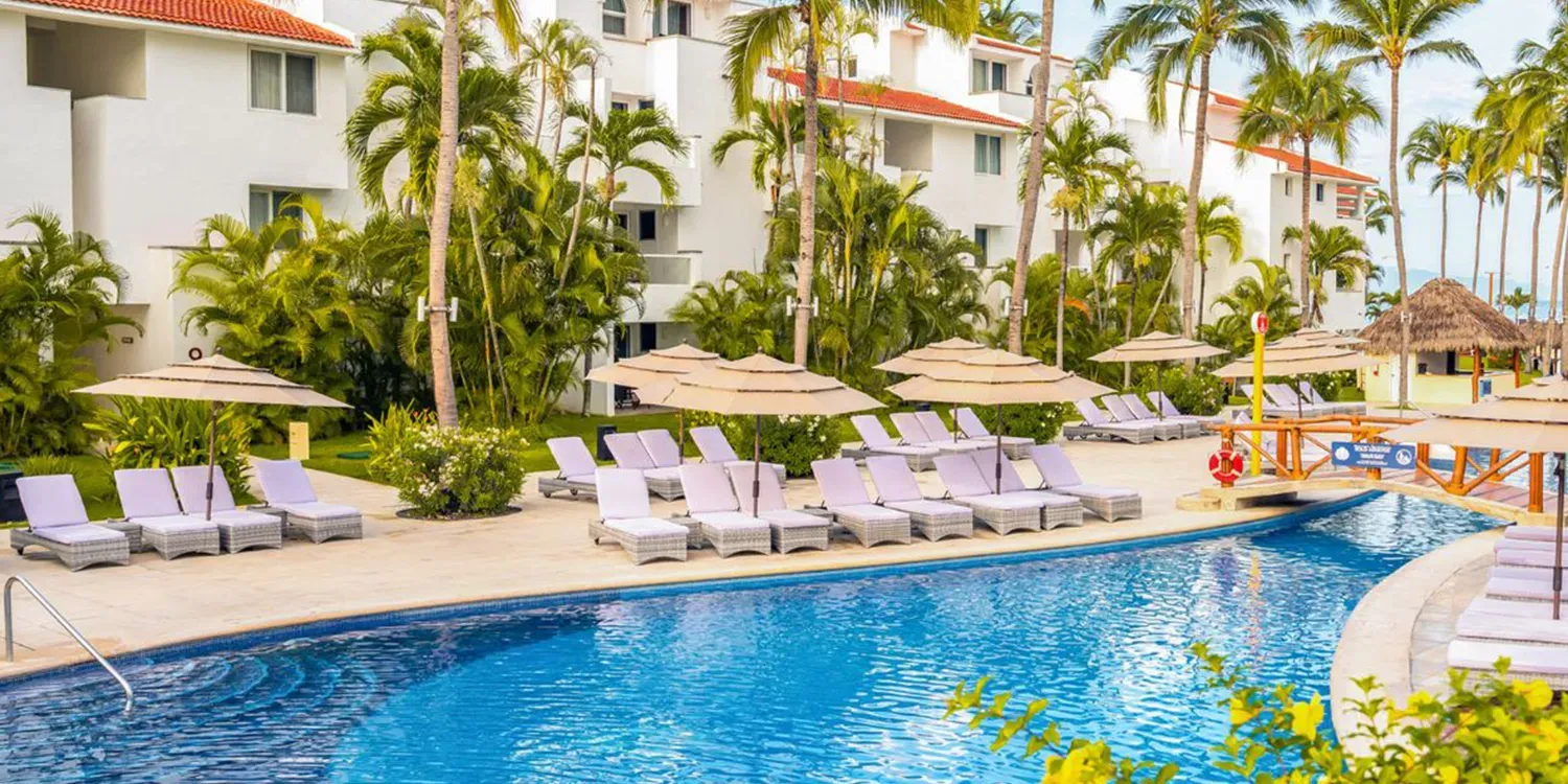 Swimming pool area with lounge chairs, umbrellas, and tropical resort buildings.