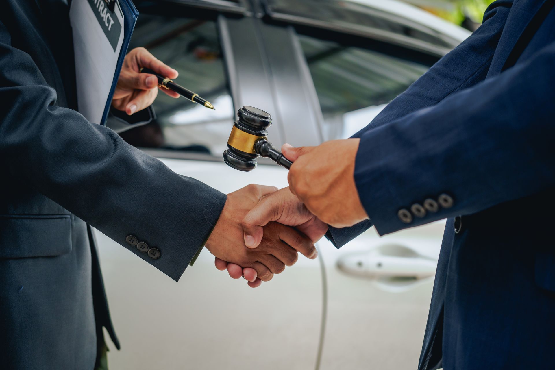 Two people shaking hands, auctioneer's gavel present, near a car.