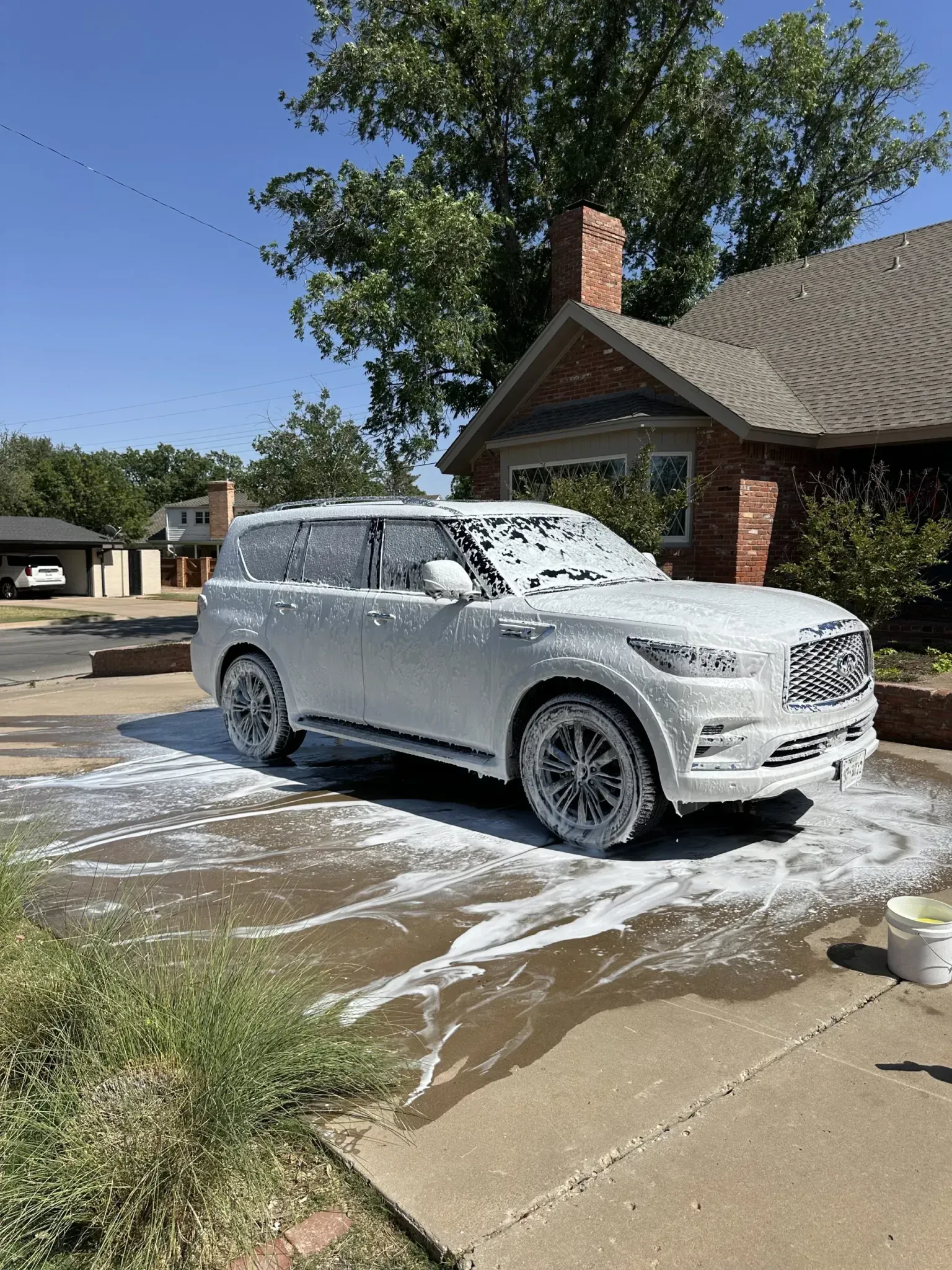 White SUV covered in soapy foam, parked on a driveway in front of a brick house.