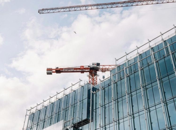 Construction of a modern glass-walled building with two cranes against a cloudy sky.