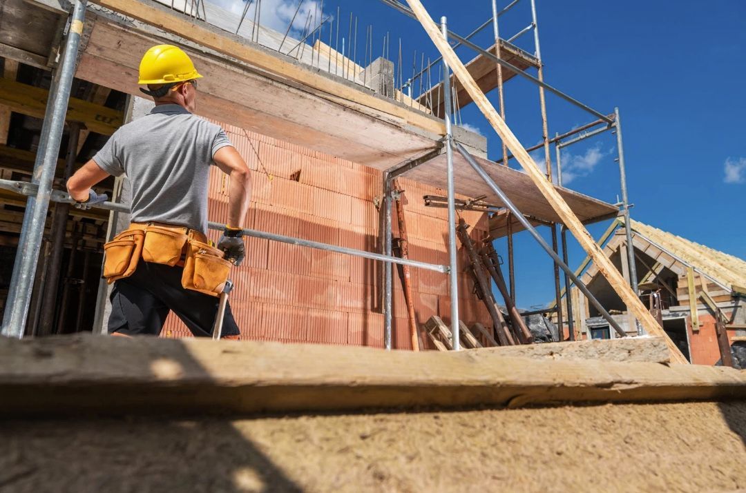 Construction worker on scaffolding, wearing a yellow hard hat and tool belt, building a brick structure on a sunny day.