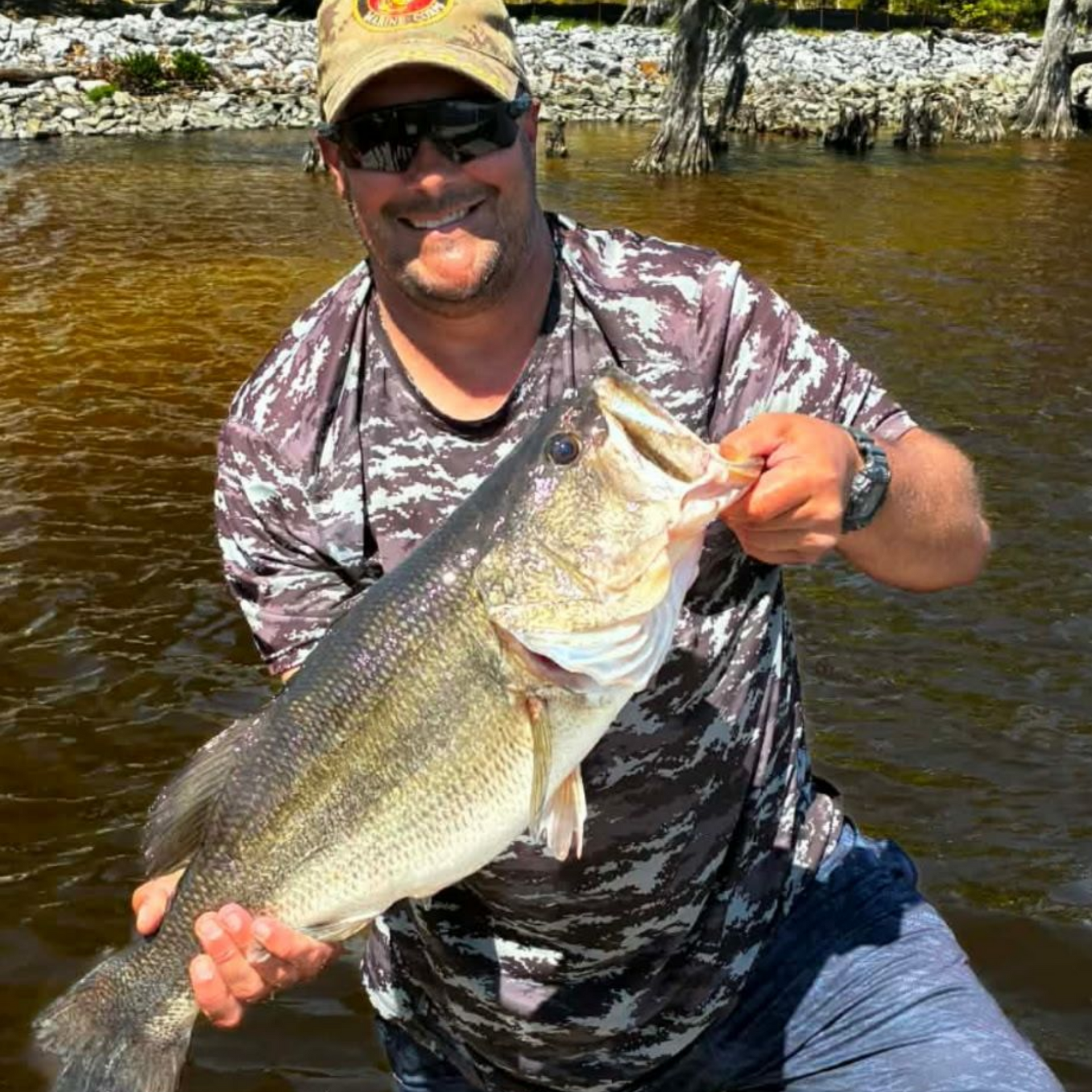 Smiling angler kneels in shallow water, holding a large bass by the shore.