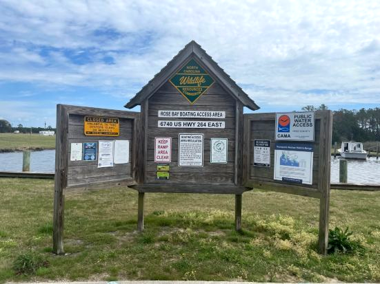 Wooden signboard with multiple notices by a waterfront under a cloudy sky