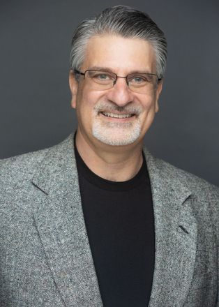 A man with glasses and a beard is wearing a black shirt and a grey jacket.