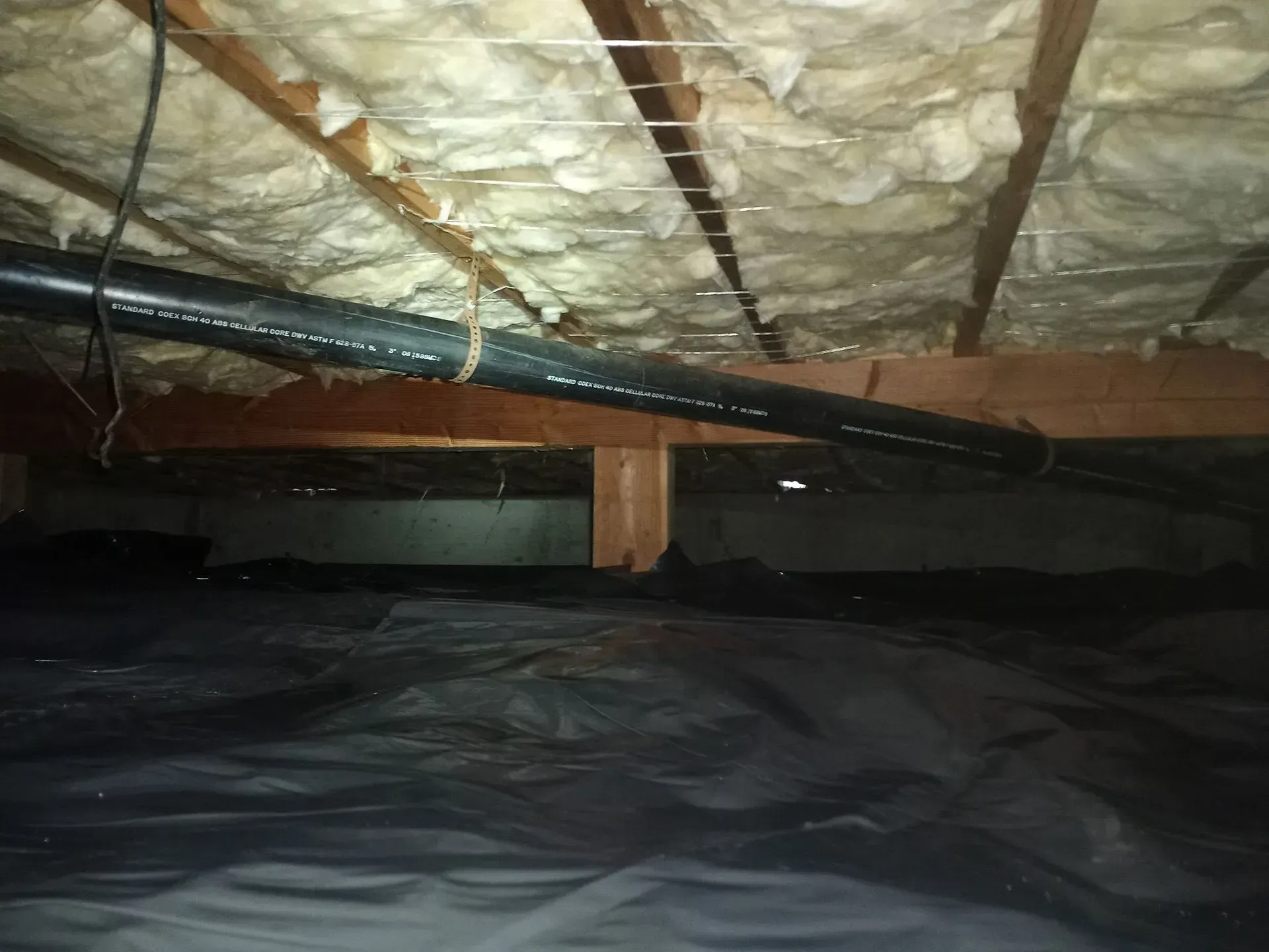 Black plastic ground cover in a crawlspace under a house, with plumbing and insulation above.