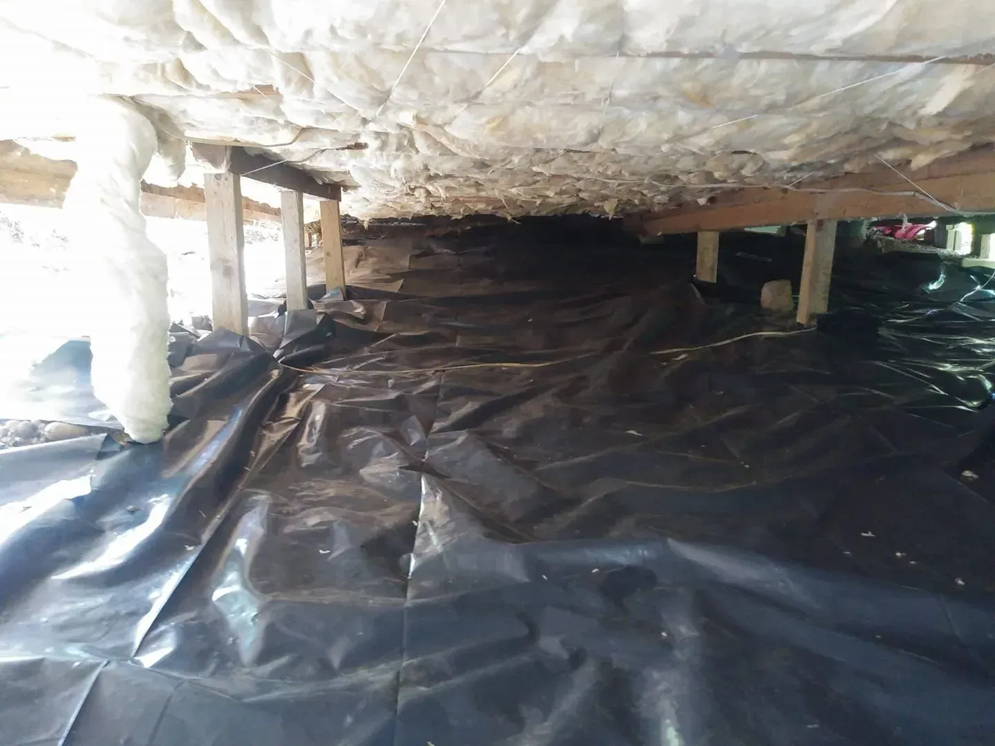 Underneath a house, a black plastic vapor barrier covers the ground. Insulation and wooden supports are visible.