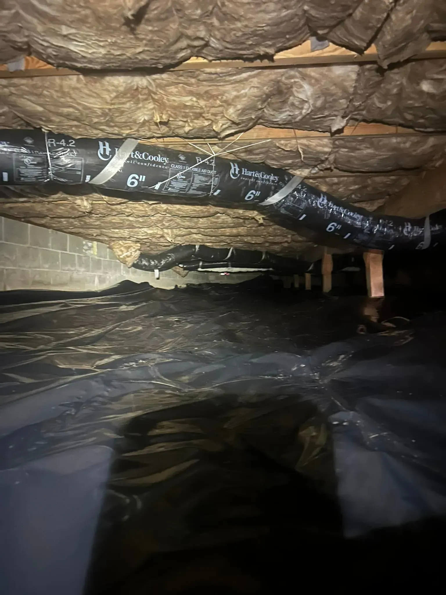Dark crawlspace with insulation, ductwork, and plastic sheeting.