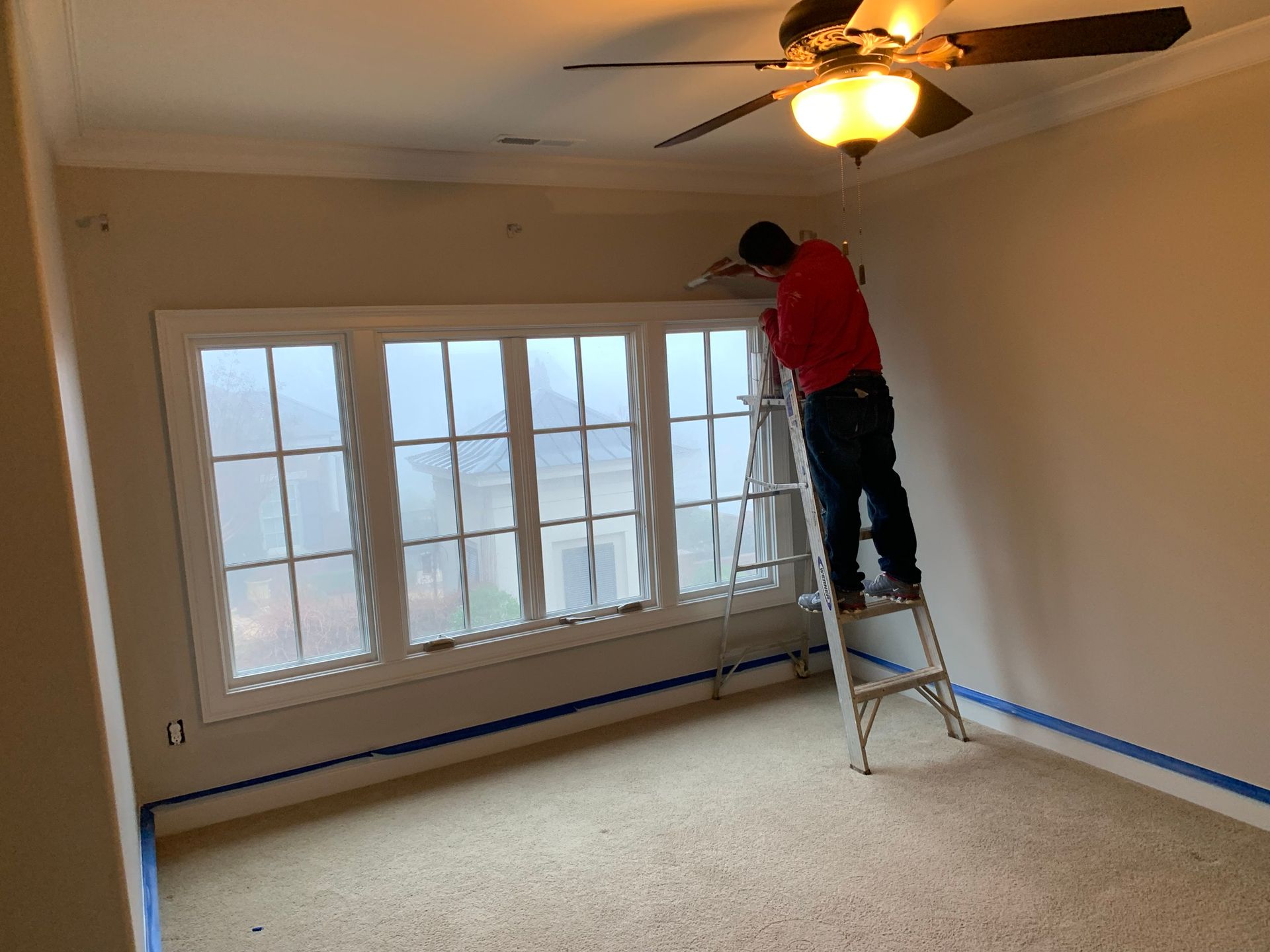 A man is standing on a ladder painting a window in a room.