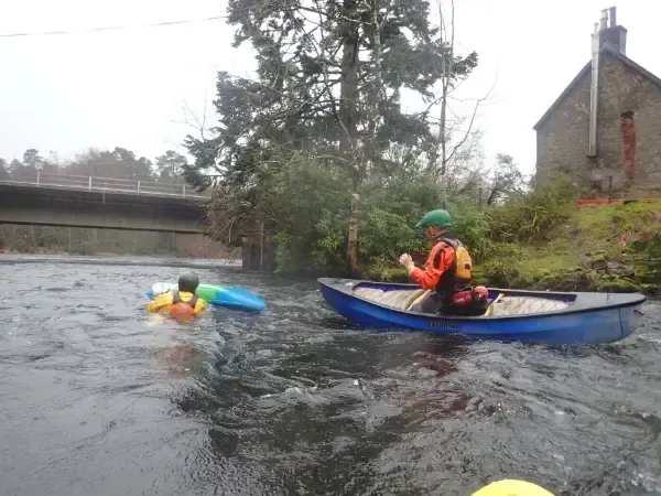 Mae dyn mewn canŵ glas yn padlo i lawr afon