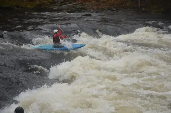 Mae person yn marchogaeth caiac i lawr afon.