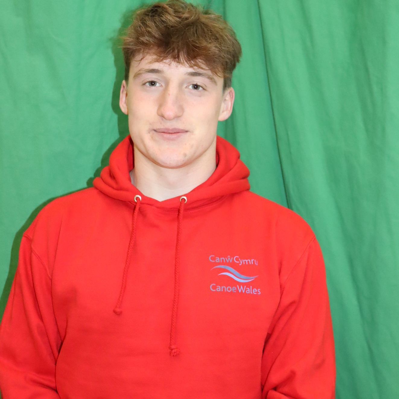 A young man wearing a red hoodie is standing in front of a green curtain.
