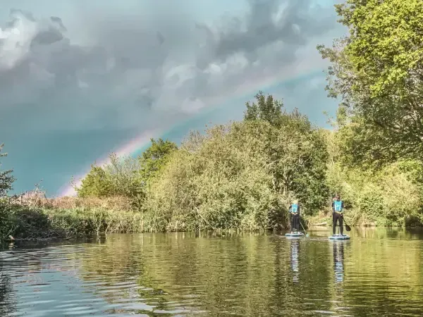 Mae dau berson yn reidio byrddau padlo ar afon o dan enfys.