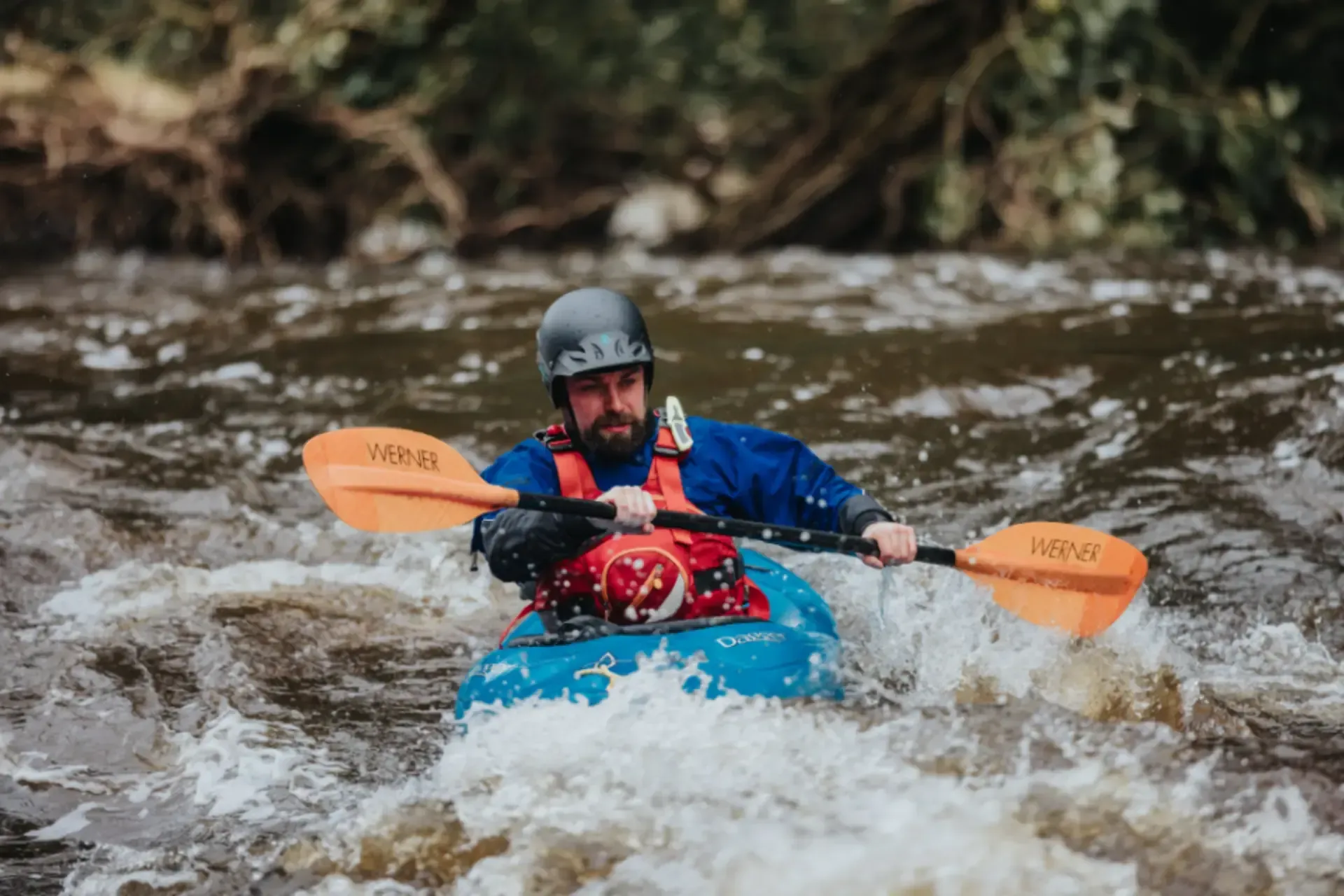 Mae dyn yn padlo caiac i lawr afon.