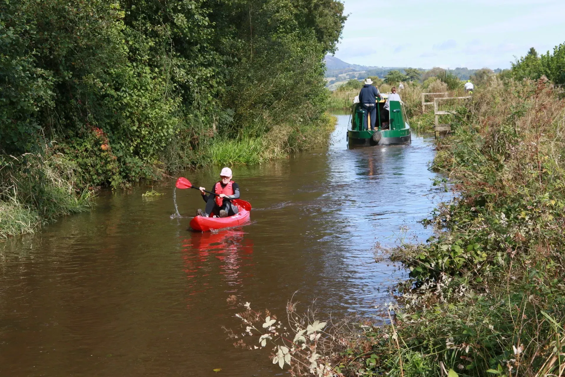 A man in a red kayak is paddling down a river next to a green boat.
