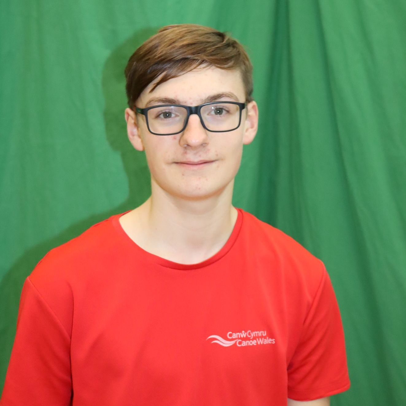 A young man wearing glasses and a red shirt stands in front of a green curtain